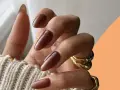 Mani con unghie autunnali chiare color cioccolato, lunghe e affusolate, adornate da anelli dorati.