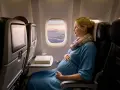 Donna incinta in aereo, con la pancia che si intravede sotto il vestito blu, guarda fuori dal finestrino le nuvole al tramonto.