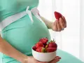 Pancia di donna incinta in abito verde acqua, con una ciotola di fragole in grembo e una fragola in mano.