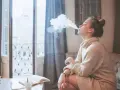 Ragazza con iqos in allattamento, fuma una nuvola di vapore mentre legge un libro sul divano.