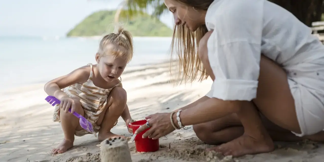 Bambina bionda e mamma giocano con secchiello e paletta sulla sabbia. Un'idea su cosa portare al mare con bambini per divertirsi.