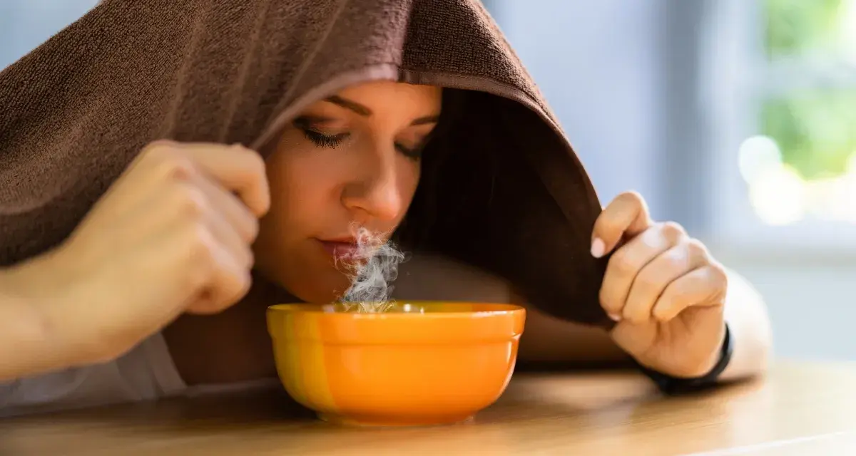 Donna inala vapore da una ciotola, usando un asciugamano per creare una sauna facciale. Un rimedio naturale per sciogliere il catarro.