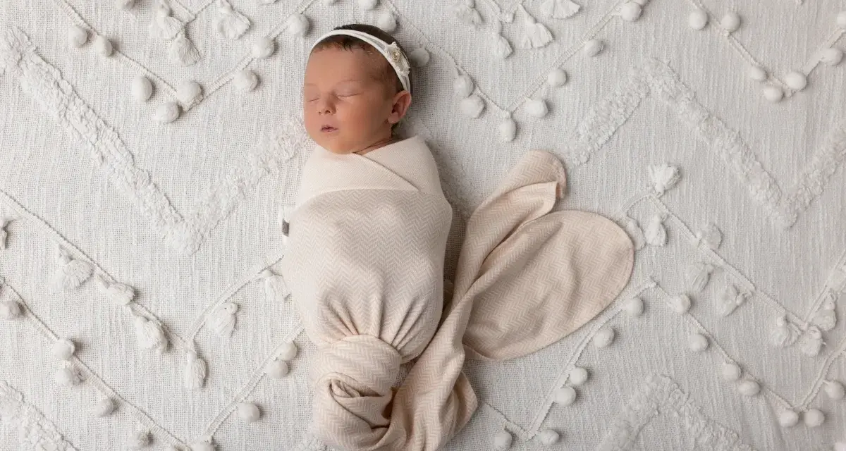 Dolce bambino in fasce dorme sereno su una coperta bianca decorata con pompon e nappine.