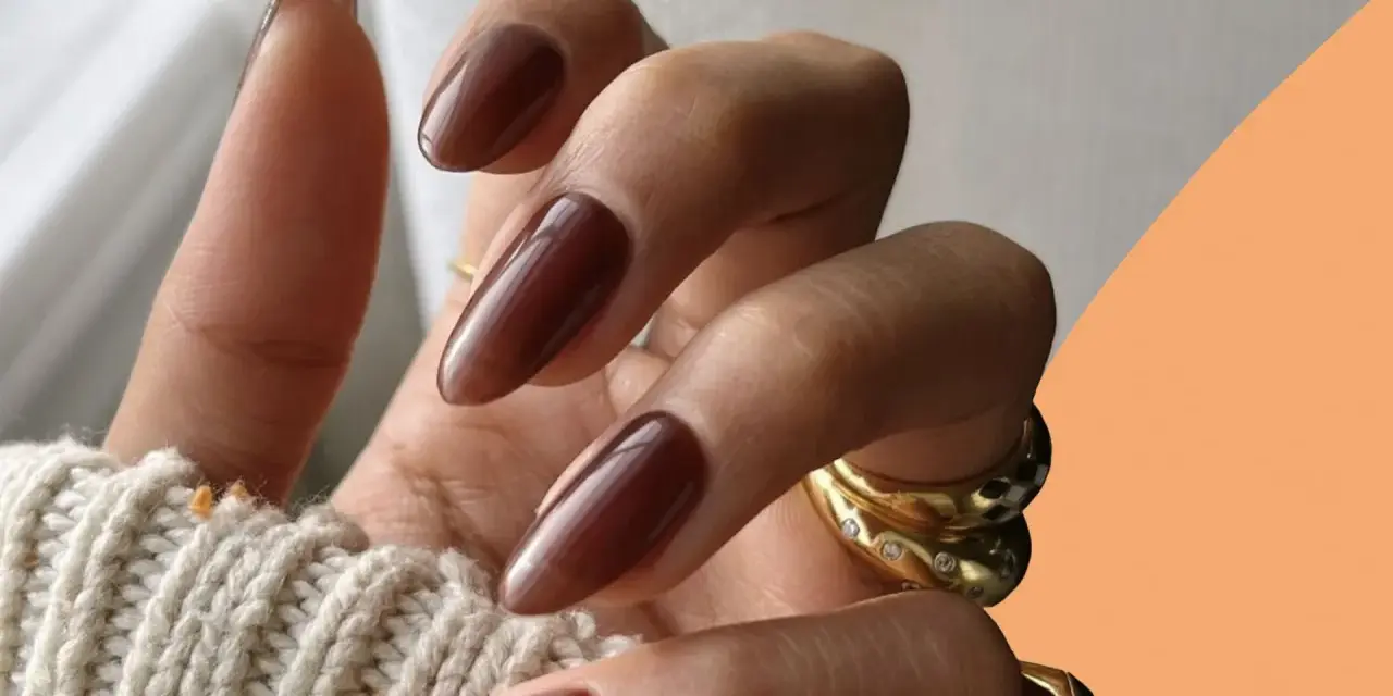 Mani con unghie autunnali chiare color cioccolato, lunghe e affusolate, adornate da anelli dorati.