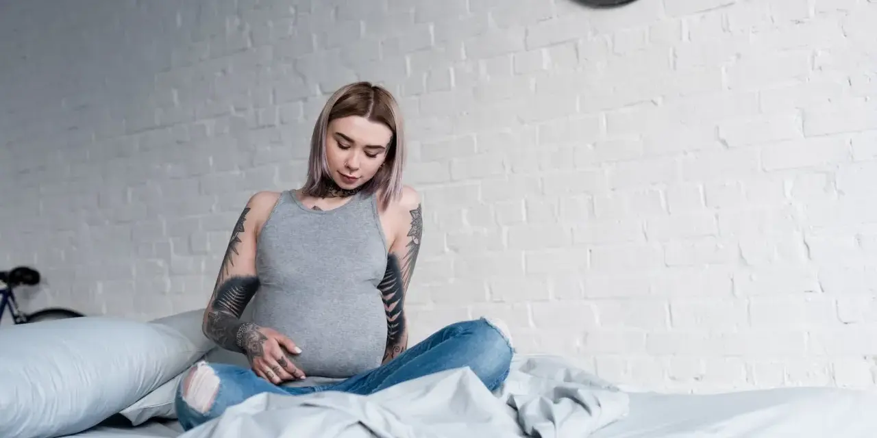 Donna incinta con tatuaggi sulle braccia e sul collo, seduta sul letto, con un pancione visibile.
