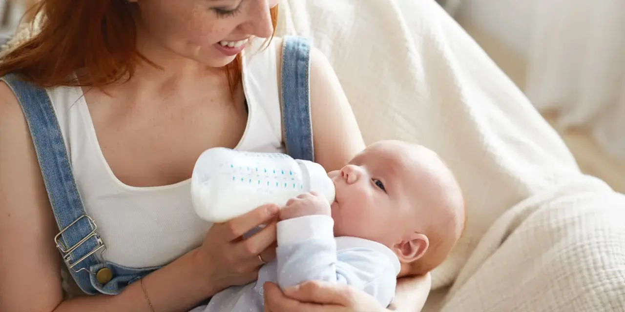Mamma con capelli rossi allatta il suo bambino con un biberon. Impara come lavare i biberon per garantire l'igiene.
