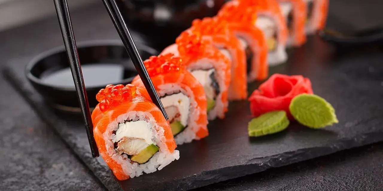 Un rotolo di sushi, con salmone e uova di pesce, viene sollevato con le bacchette. Accanto, zenzero marinato e wasabi. Un vero piacere per il palato, un sushi in allattamento di sapori.