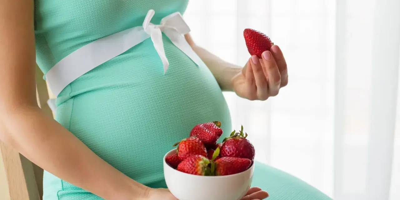 Pancia di donna incinta in abito verde acqua, con una ciotola di fragole in grembo e una fragola in mano.