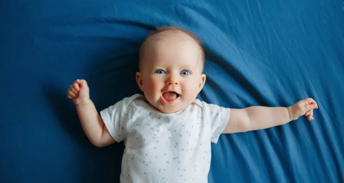 Un bimbo sorridente, con occhi azzurri e braccia aperte, celebra il suo scatto di crescita di 4 mesi su un telo blu.