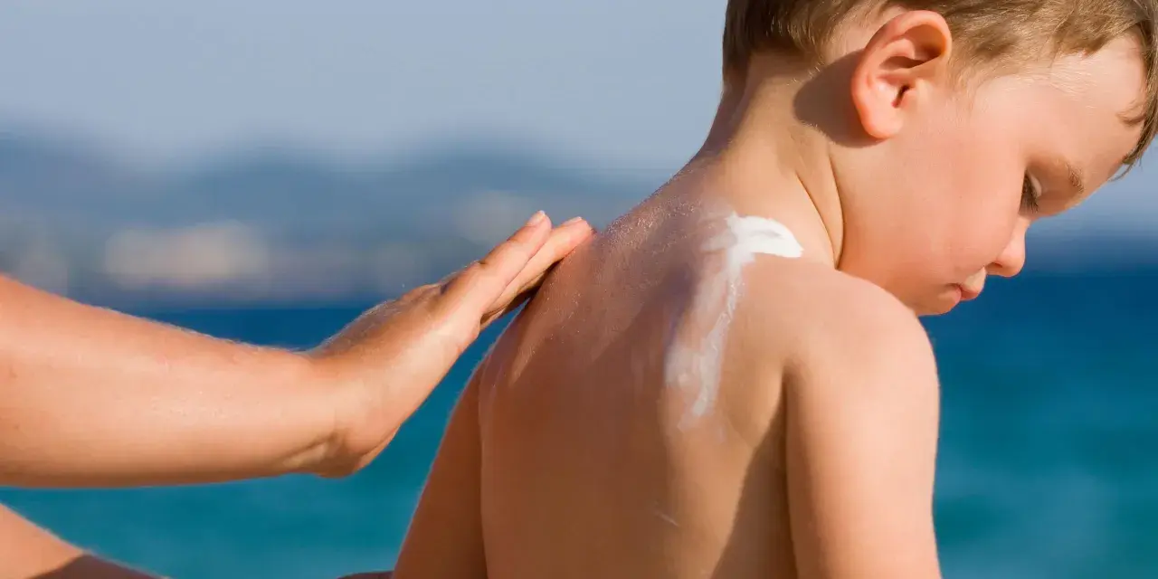 Mano applica crema solare neonato 2 mesi sulla schiena di un bimbo, con il mare sullo sfondo.