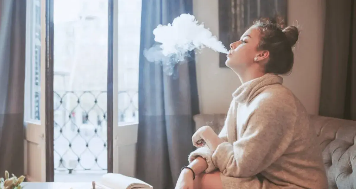 Ragazza con iqos in allattamento, fuma una nuvola di vapore mentre legge un libro sul divano.