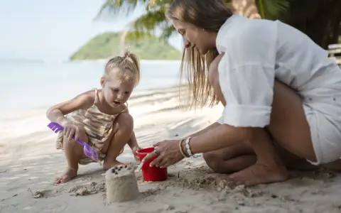 Bambina bionda e mamma giocano con secchiello e paletta sulla sabbia. Un'idea su cosa portare al mare con bambini per divertirsi.