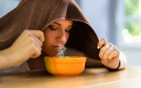 Donna inala vapore da una ciotola, usando un asciugamano per creare una sauna facciale. Un rimedio naturale per sciogliere il catarro.
