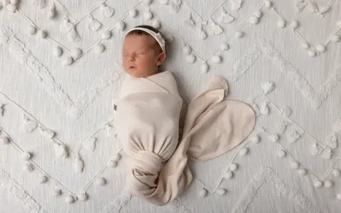 Dolce bambino in fasce dorme sereno su una coperta bianca decorata con pompon e nappine.