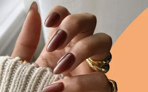 Mani con unghie autunnali chiare color cioccolato, lunghe e affusolate, adornate da anelli dorati.