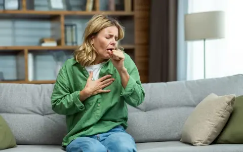 Donna tossisce sul divano, con una mano sul petto, cercando sollievo per la bronchite con un antibiotico.