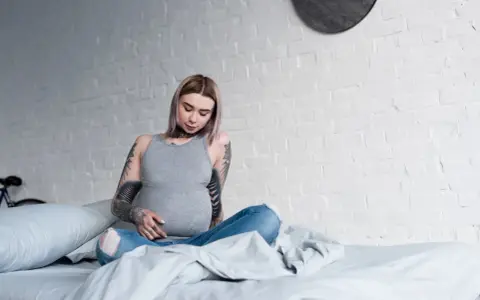 Donna incinta con tatuaggi sulle braccia e sul collo, seduta sul letto, con un pancione visibile.