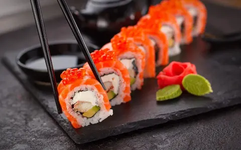 Un rotolo di sushi, con salmone e uova di pesce, viene sollevato con le bacchette. Accanto, zenzero marinato e wasabi. Un vero piacere per il palato, un sushi in allattamento di sapori.