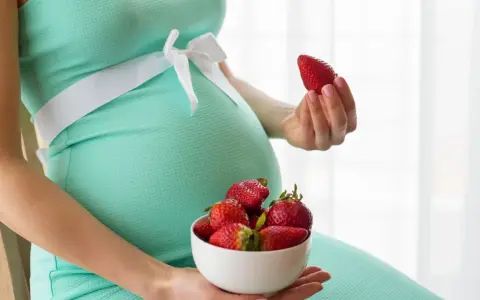 Pancia di donna incinta in abito verde acqua, con una ciotola di fragole in grembo e una fragola in mano.