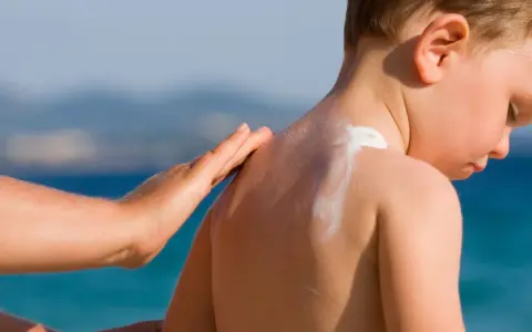 Mano applica crema solare neonato 2 mesi sulla schiena di un bimbo, con il mare sullo sfondo.