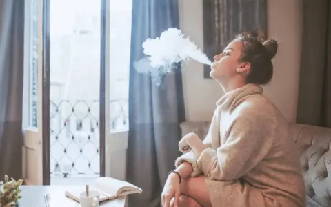 Ragazza con iqos in allattamento, fuma una nuvola di vapore mentre legge un libro sul divano.