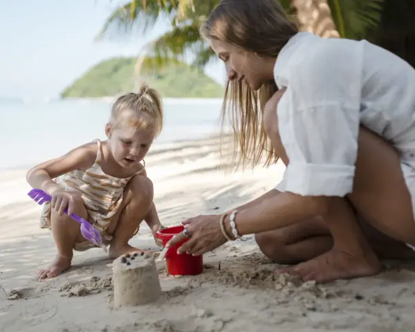 Bambina bionda e mamma giocano con secchiello e paletta sulla sabbia. Un'idea su cosa portare al mare con bambini per divertirsi.