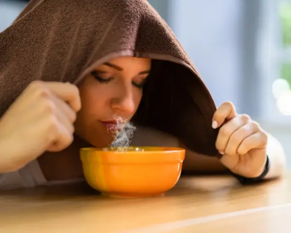 Donna inala vapore da una ciotola, usando un asciugamano per creare una sauna facciale. Un rimedio naturale per sciogliere il catarro.