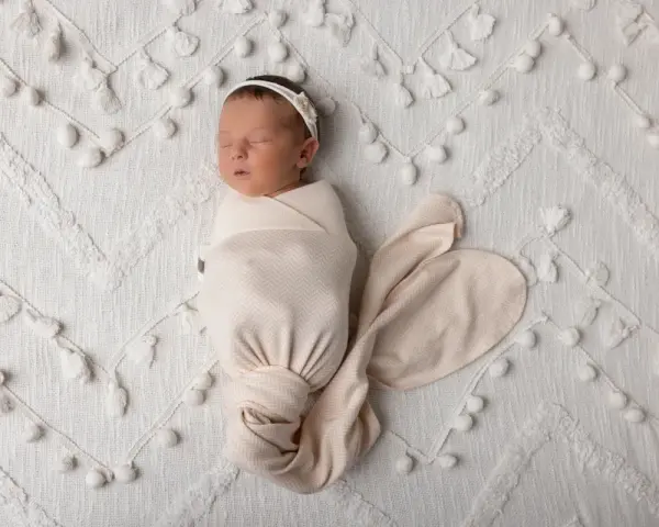 Dolce bambino in fasce dorme sereno su una coperta bianca decorata con pompon e nappine.