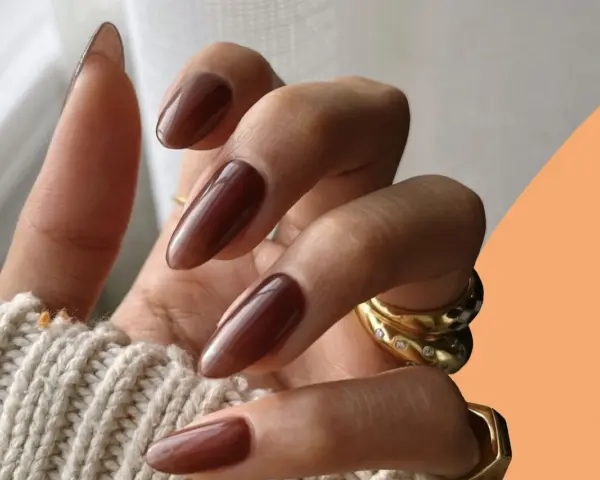 Mani con unghie autunnali chiare color cioccolato, lunghe e affusolate, adornate da anelli dorati.