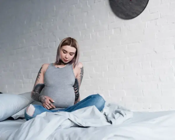 Donna incinta con tatuaggi sulle braccia e sul collo, seduta sul letto, con un pancione visibile.