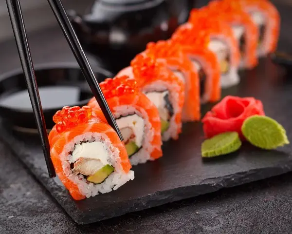 Un rotolo di sushi, con salmone e uova di pesce, viene sollevato con le bacchette. Accanto, zenzero marinato e wasabi. Un vero piacere per il palato, un sushi in allattamento di sapori.