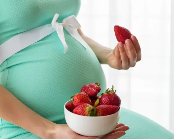 Pancia di donna incinta in abito verde acqua, con una ciotola di fragole in grembo e una fragola in mano.