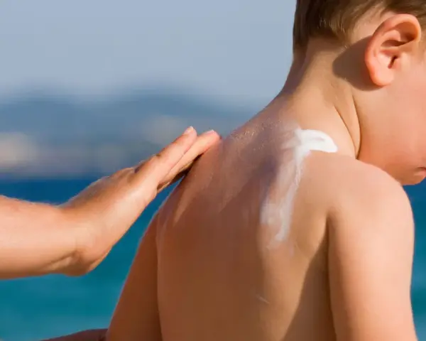 Mano applica crema solare neonato 2 mesi sulla schiena di un bimbo, con il mare sullo sfondo.