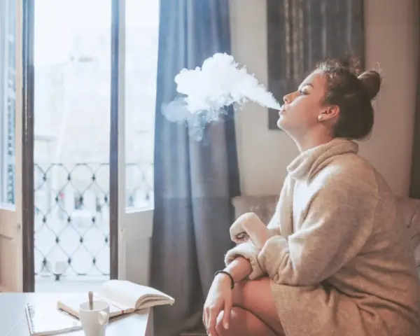 Ragazza con iqos in allattamento, fuma una nuvola di vapore mentre legge un libro sul divano.