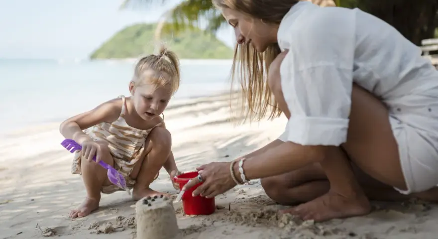 Bambina bionda e mamma giocano con secchiello e paletta sulla sabbia. Un'idea su cosa portare al mare con bambini per divertirsi.