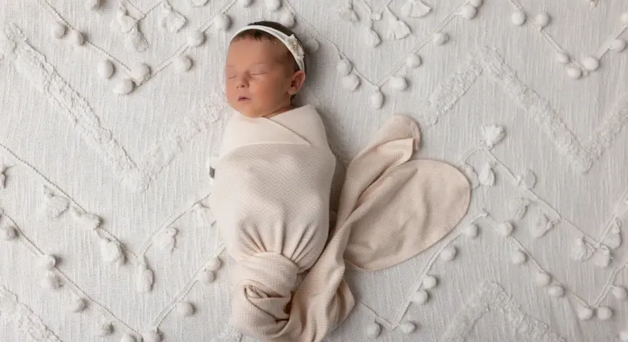 Dolce bambino in fasce dorme sereno su una coperta bianca decorata con pompon e nappine.