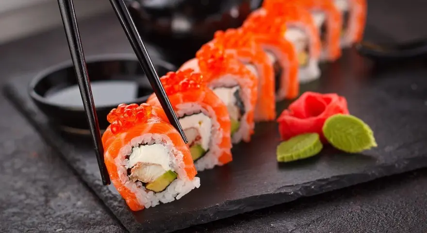 Un rotolo di sushi, con salmone e uova di pesce, viene sollevato con le bacchette. Accanto, zenzero marinato e wasabi. Un vero piacere per il palato, un sushi in allattamento di sapori.
