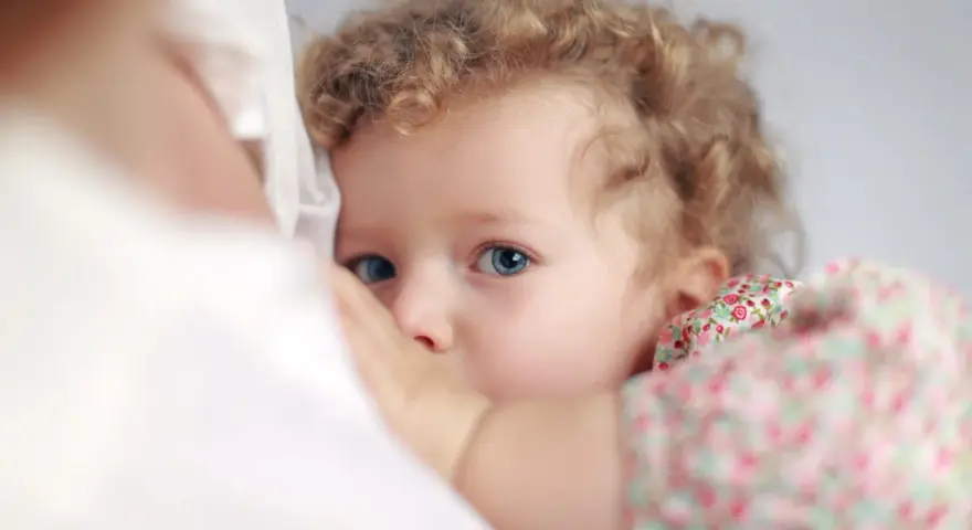 Bambina con occhi azzurri e capelli ricci, stretta al petto della madre. Un'immagine che evoca il legame profondo, ma anche le possibili conseguenze psicologiche dell'allattamento prolungato.
