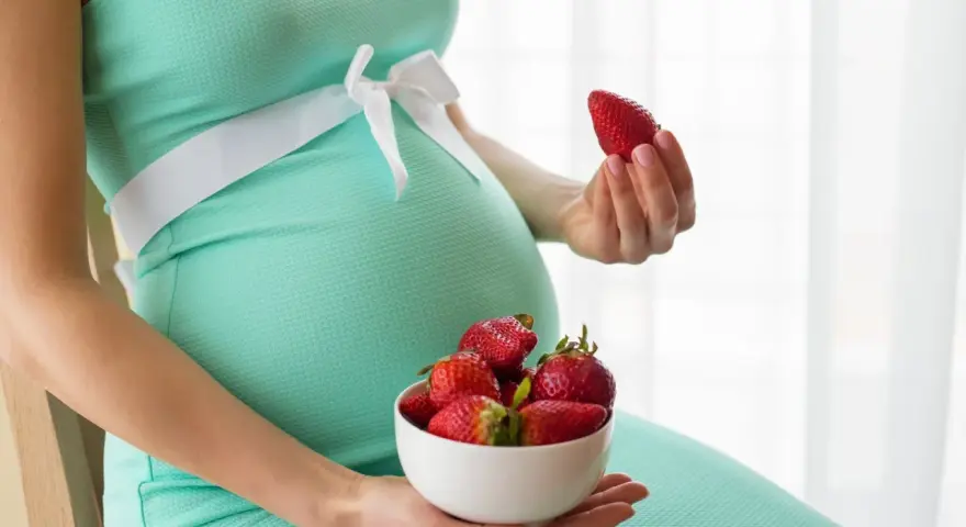 Pancia di donna incinta in abito verde acqua, con una ciotola di fragole in grembo e una fragola in mano.