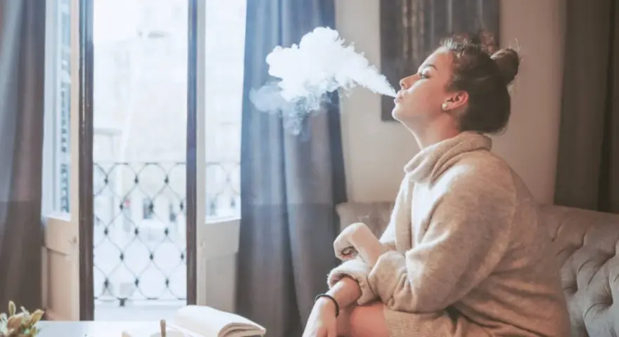 Ragazza con iqos in allattamento, fuma una nuvola di vapore mentre legge un libro sul divano.