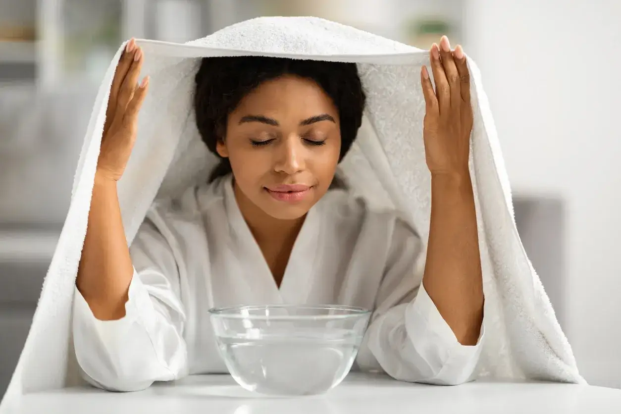 Donna con gli occhi chiusi inala vapore da una ciotola d'acqua, usando un asciugamano come tenda. Un rimedio naturale per i fumenti per catarro.