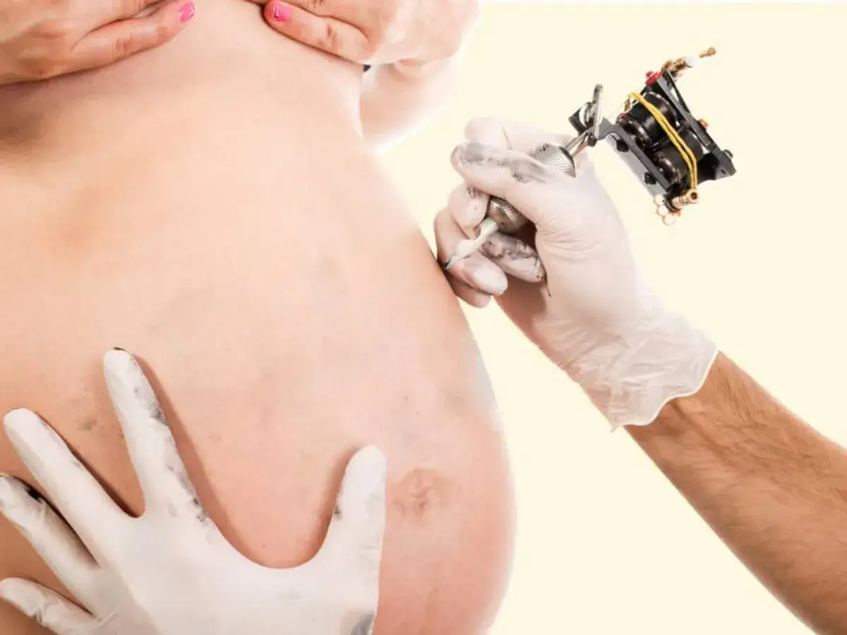 Mano guantata con macchinetta per tatuaggi vicino a pancia di donna incinta. Tatuaggi in gravidanza, un ricordo indelebile.
