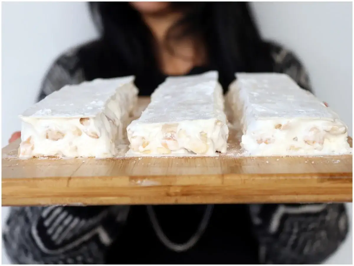 Tre blocchi di torrone bianco con mandorle, spolverati di zucchero a velo, su un tagliere di legno. Un dolce perfetto per chi aspetta un bambino.