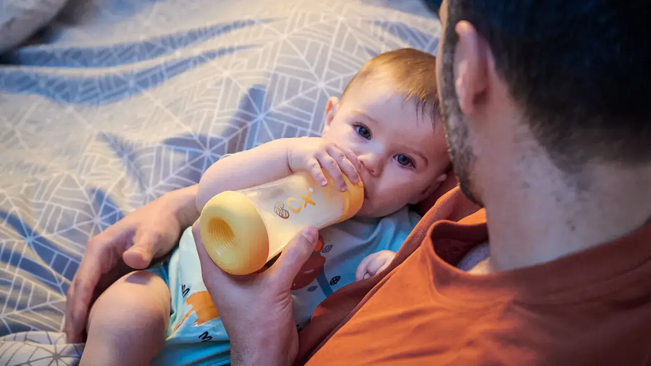 Un padre tiene un biber&oacute;n per dare da mangiare al suo neonato, un momento tenero su un letto con lenzuola a fantasia.