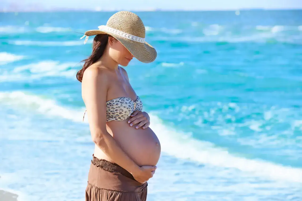 Donna incinta con cappello di paglia sulla spiaggia, accarezza il pancione. Il caldo in gravidanza si affronta meglio con una brezza marina.