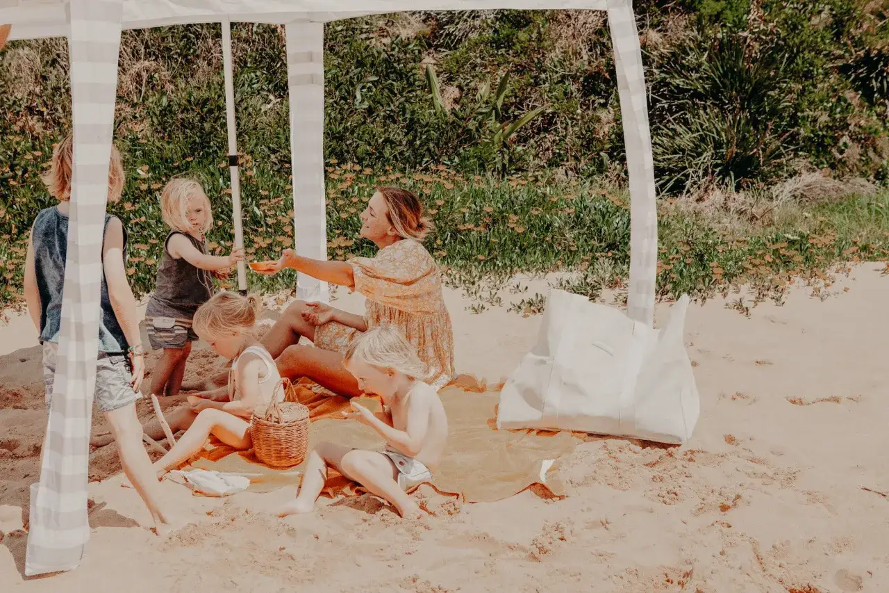 Famiglia in spiaggia sotto una tenda, con bambini che mangiano frutta. Un esempio di cosa portare al mare con bambini per un picnic felice.