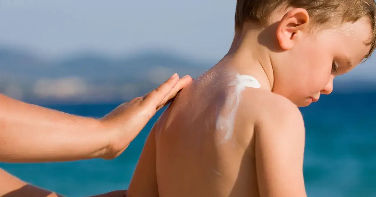 Mano applica crema solare neonato 2 mesi sulla schiena di un bimbo, con il mare sullo sfondo.